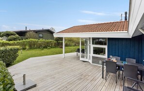 Eine Holzterrasse mit überdachter Veranda, einem Tisch mit Stühlen und Blick auf einen Garten sowie angrenzende Gebäude.