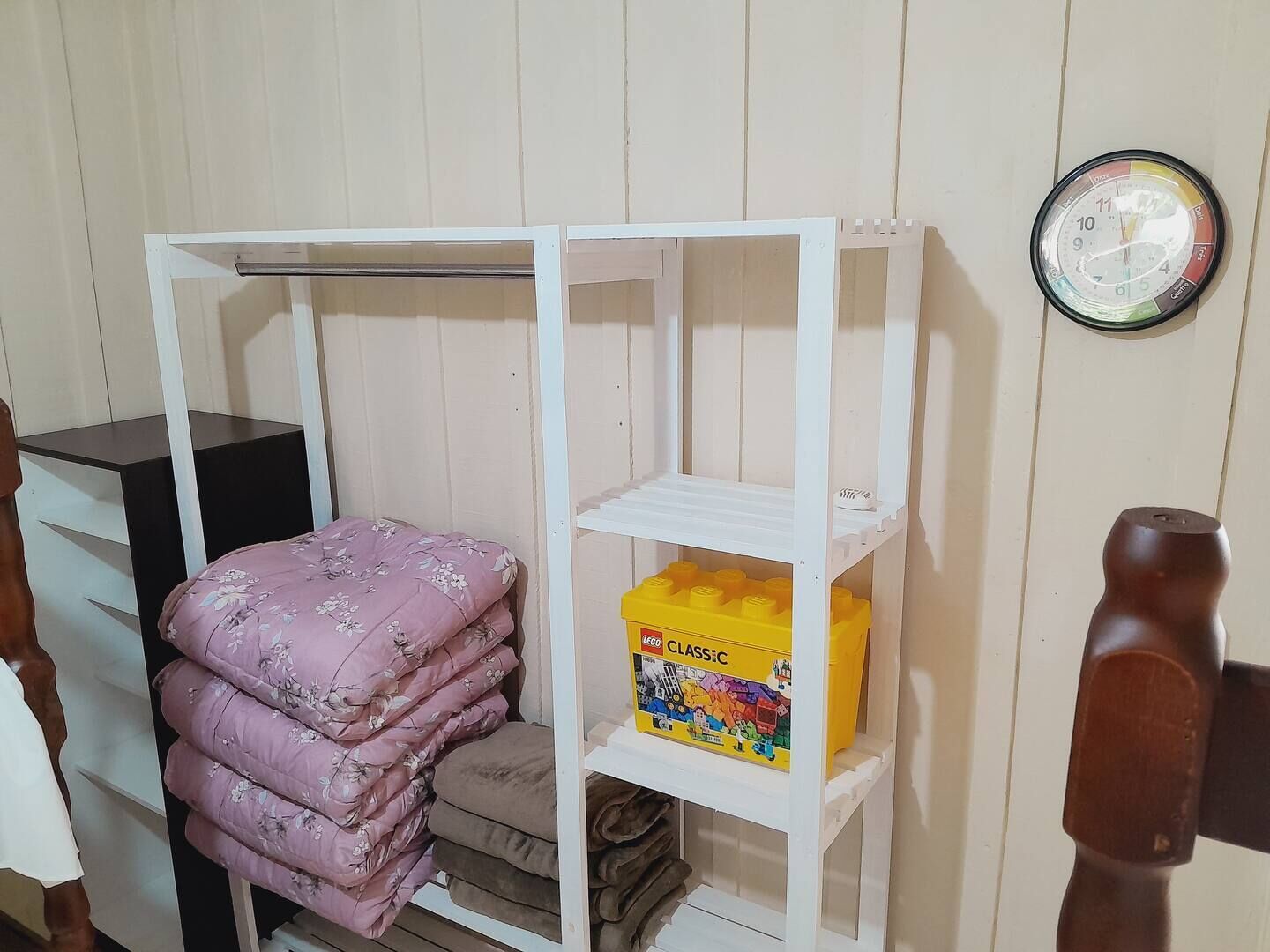 Um quarto com uma cama, um guarda-roupa, uma prateleira com cobertores dobrados, uma caixa de LEGO e um relógio de parede.