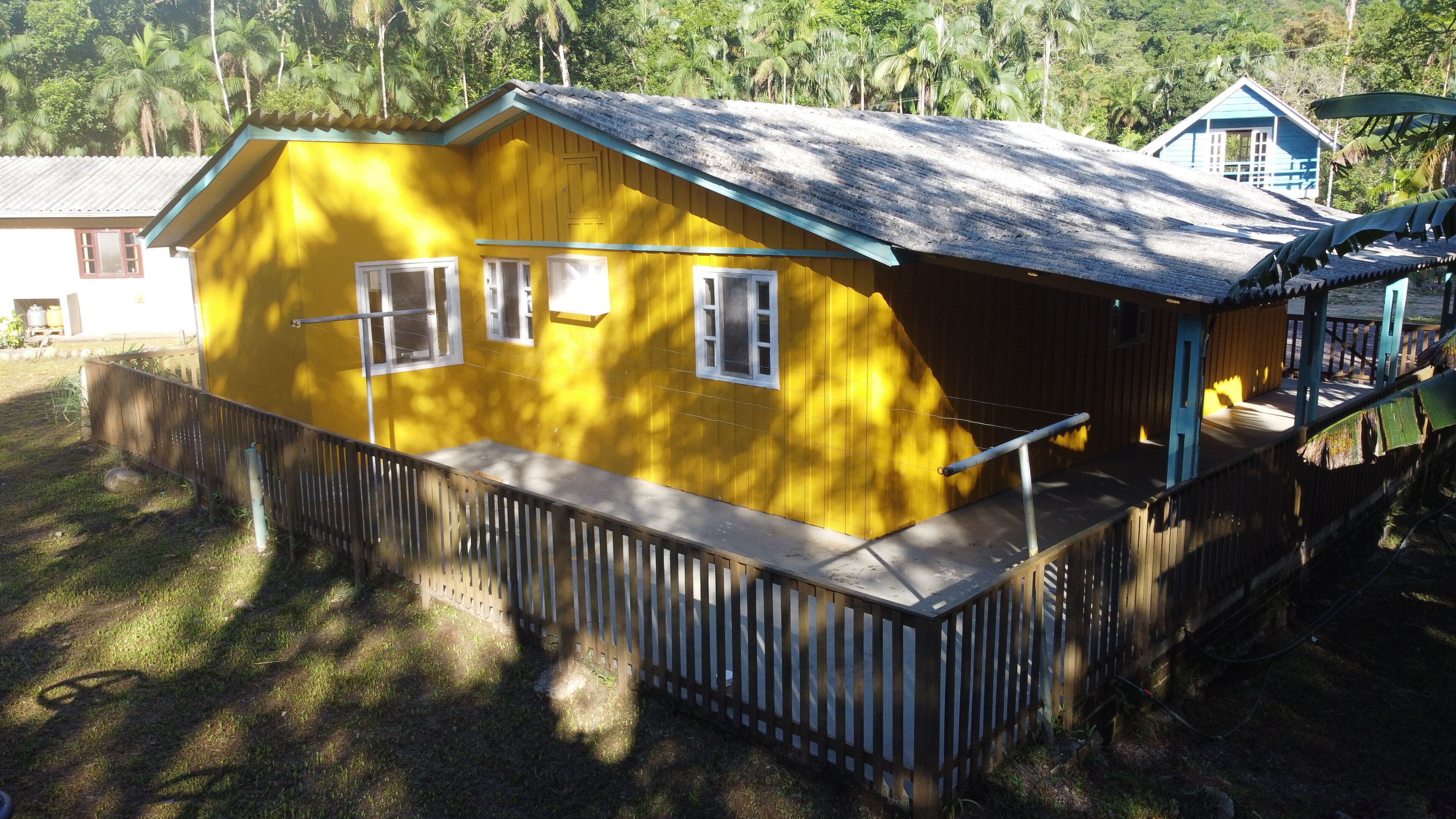 A yellow house with a porch, a fenced yard, and a blue building in the background.