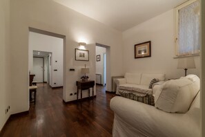 A spacious living room with a wooden floor, a white sofa, and a lamp. There is a hallway leading to another room.