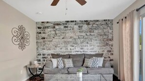A living room with a grey sofa, patterned cushions, a glass coffee table, a decorative wall piece, and a brick wall backdrop. - Experience Scottsdale Charm: Cozy Retreat, Heated Pool, Family-Friendly Fun! (Scottsdale)
