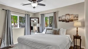 A bedroom with a large bed, a ceiling fan, a window with curtains, a bedside table with a lamp, and wall art. - Experience Scottsdale Charm: Cozy Retreat, Heated Pool, Family-Friendly Fun! (Scottsdale)