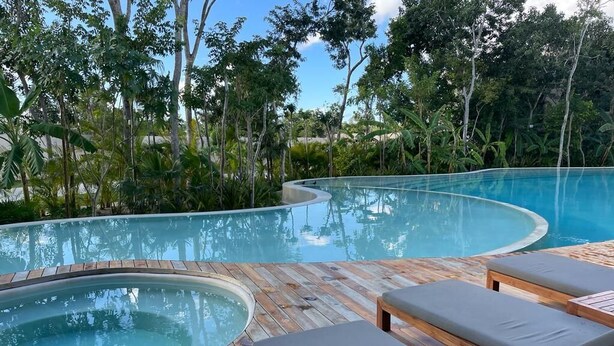 A curved infinity pool with wooden deck and lounge chairs, surrounded by tropical vegetation.