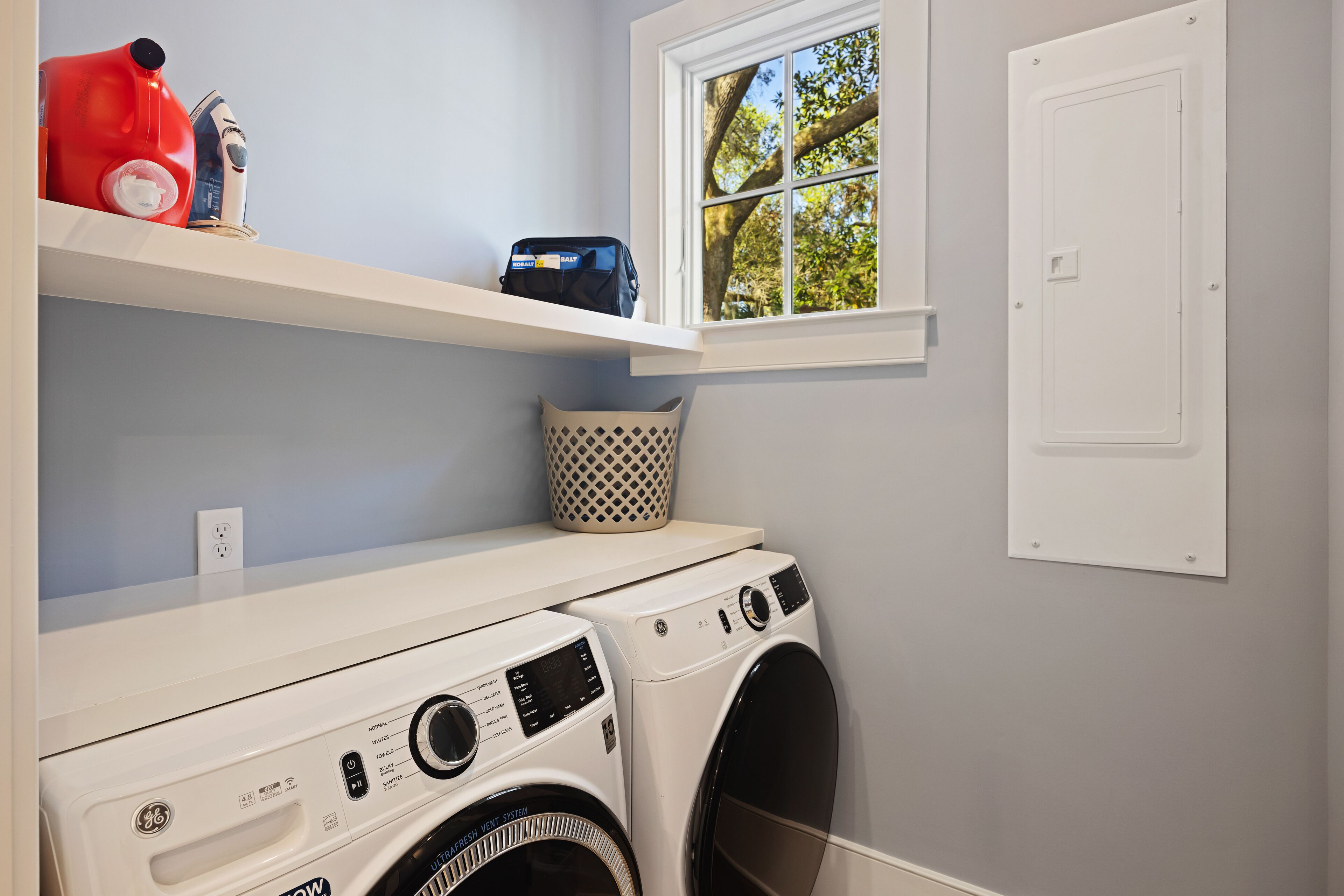 A laundry area with two front-loading washing machines, a shelf with a red water container, a blue bag, and a patterned basket.