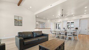 Uma sala de estar ampla com um sofá de couro escuro, uma mesa de centro de madeira e uma área de jantar com cadeiras brancas e uma mesa de madeira.