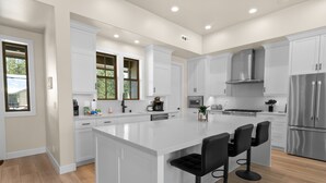 Cozinha moderna com armários brancos, uma ilha grande com banquetas, eletrodomésticos de aço inoxidável e vista para o exterior.