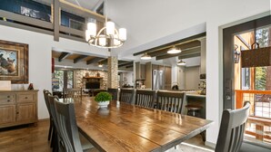 A spacious dining area with a wooden table and chairs, a chandelier, and a stone fireplace. - Evergreen - Luxury 3BR | En-Suite, Hot Tub, Game Room (Big Bear Lake)