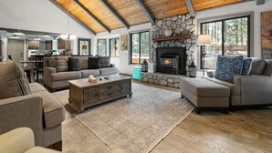 A spacious living room with a fireplace, wooden ceiling beams, and large windows. - Evergreen - Luxury 3BR | En-Suite, Hot Tub, Game Room (Big Bear Lake)