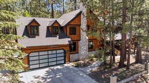 Une maison moderne à deux étages, dotée d’une façade en pierre, d’un grand garage et entourée de grands pins.