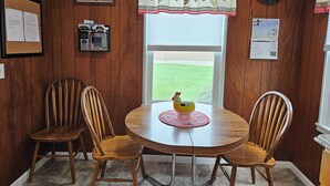 Comedor con una mesa redonda de madera, dos sillas de madera, un tablón de anuncios, un reloj de pared y una ventana con vistas a un campo verde.