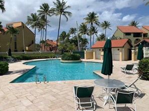 A swimming pool with lounge chairs and umbrellas, surrounded by palm trees and buildings.