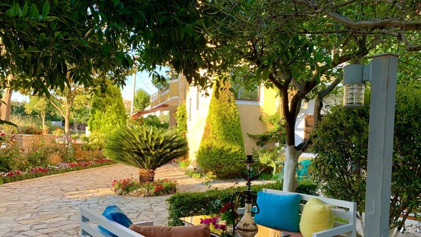 An outdoor seating area with white benches and cushions, surrounded by a garden with various plants and a decorative fountain. - Flower Garden Villa (Ksamil)