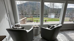 Een kamer met twee grijze fauteuils, een kleine ronde tafel en uitzicht op een terras en buitenruimte met een tuinhuis en een picknicktafel.