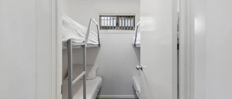 Ein schmales Gang mit Etagenbetten an beiden Seiten, einem kleinen Fenster mit Gitter und einer Tür am Ende.