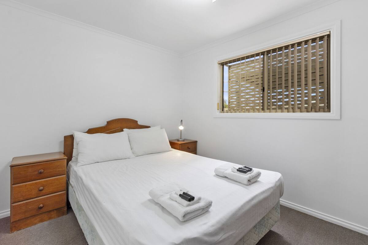 A neatly made bed with white linens, a wooden nightstand with drawers, a bedside lamp, and a window with wooden blinds.