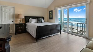 Une chambre à coucher comprenant un lit, une table de chevet, une commode, un ventilateur de plafond et une grande fenêtre donnant sur l’océan.