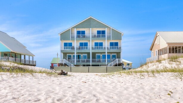 Une maison de plage à plusieurs étages, dotée d’un balcon à chaque niveau, située sur une plage de sable fin, sous un ciel dégagé.