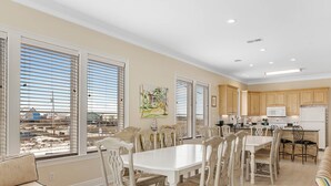 A spacious dining area with a large table and chairs, a kitchen with wooden cabinets, and large windows with blinds. - Beach Castle, West. 8BR. Sleeps 27. Large Pool! Great decks,, Beachfront. Value! (Gulf Shores)