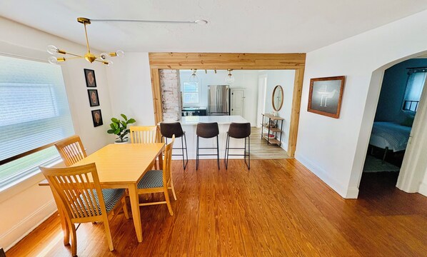 Een eetgedeelte met een houten tafel en stoelen, een keuken op de achtergrond en een gang die naar een andere kamer leidt.