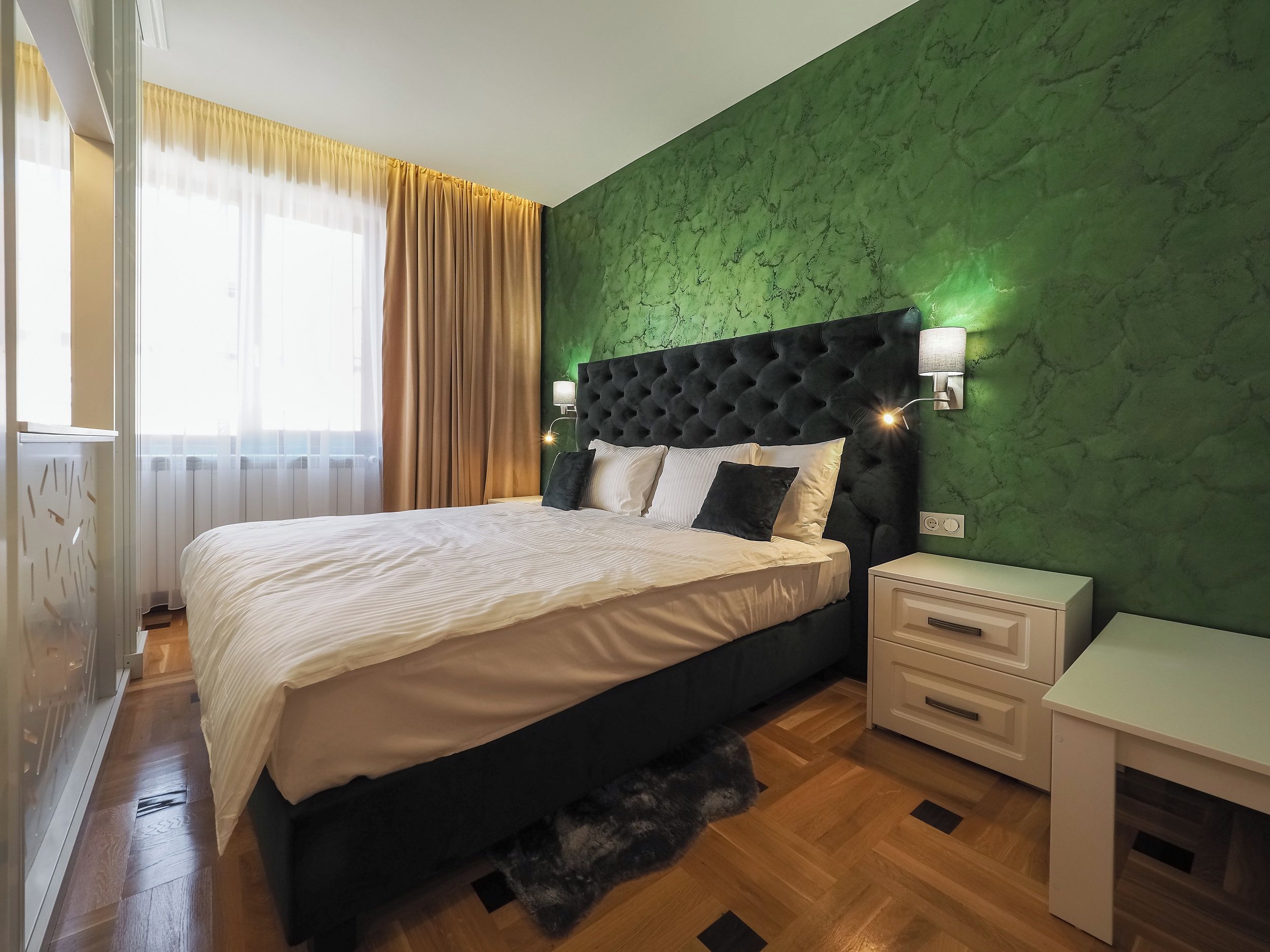 Un dormitorio con cama, mesitas de noche, una mesita auxiliar, una pared verde con textura y una ventana con cortinas.
