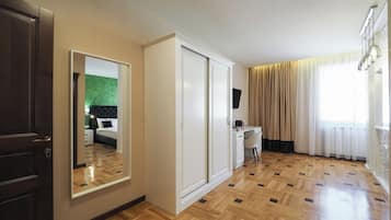 A hotel room with a wooden parquet floor, a bed with a green headboard, a desk with a chair, a wardrobe, and a large mirror.