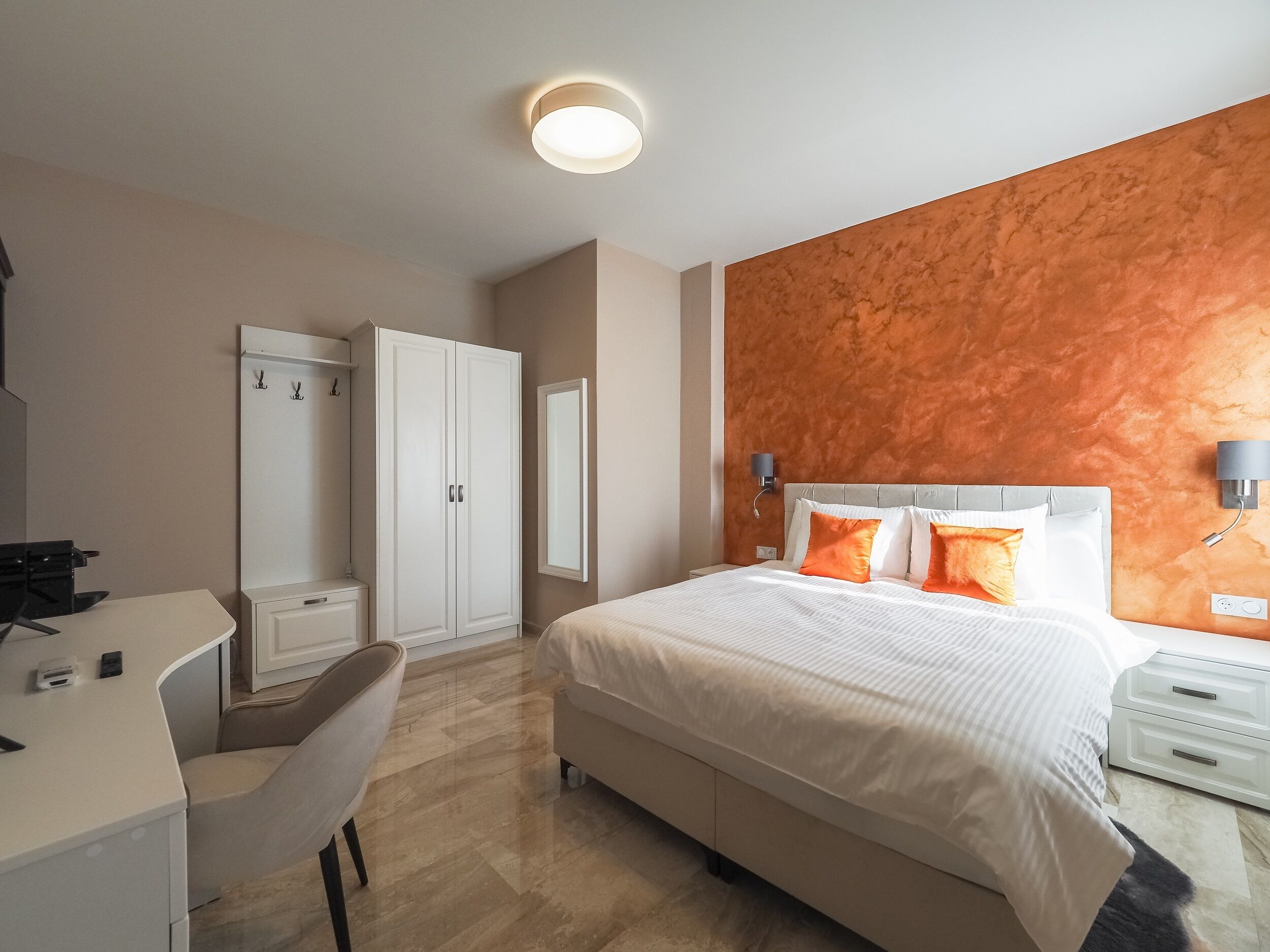 A hotel room with a bed, a desk, a chair, a wardrobe, and a wall-mounted lamp.