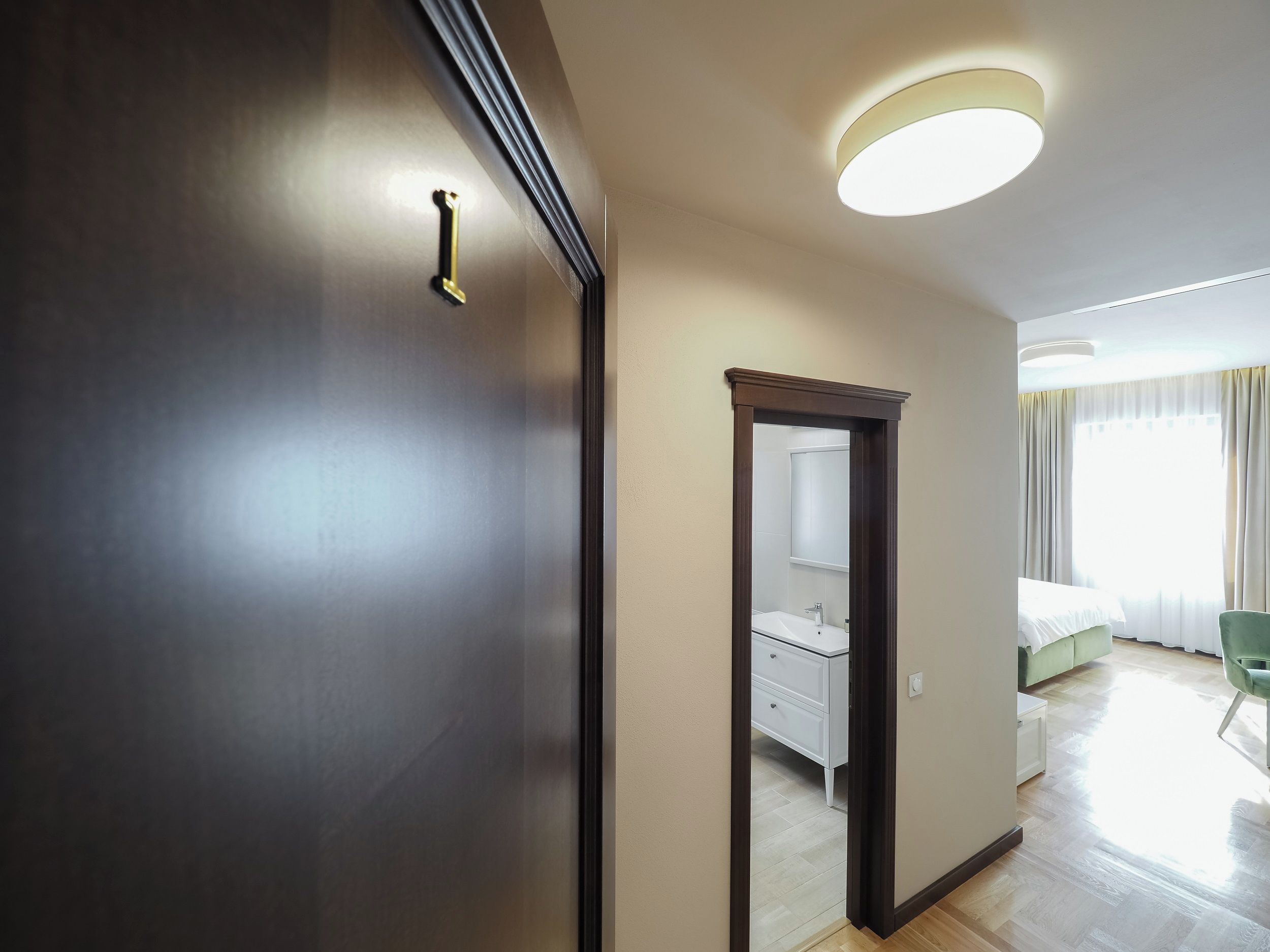 Habitación de hotel con un armario de madera, un espejo, una cama, una silla verde y un baño con un lavabo y mueble blancos.