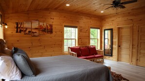 A wooden cabin bedroom with a bed, a red sofa, and a ceiling fan. - Newly Built Home: Perfectly Located Near Loon Mountain & Downtown Lincoln, NH! (Lincoln)