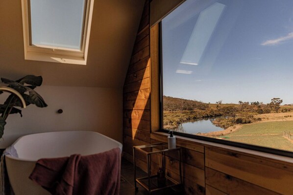 A bathroom with a bathtub, a window with a view of a lake and trees, and a wooden wall. - Swan River Sanctuary (Swansea)
