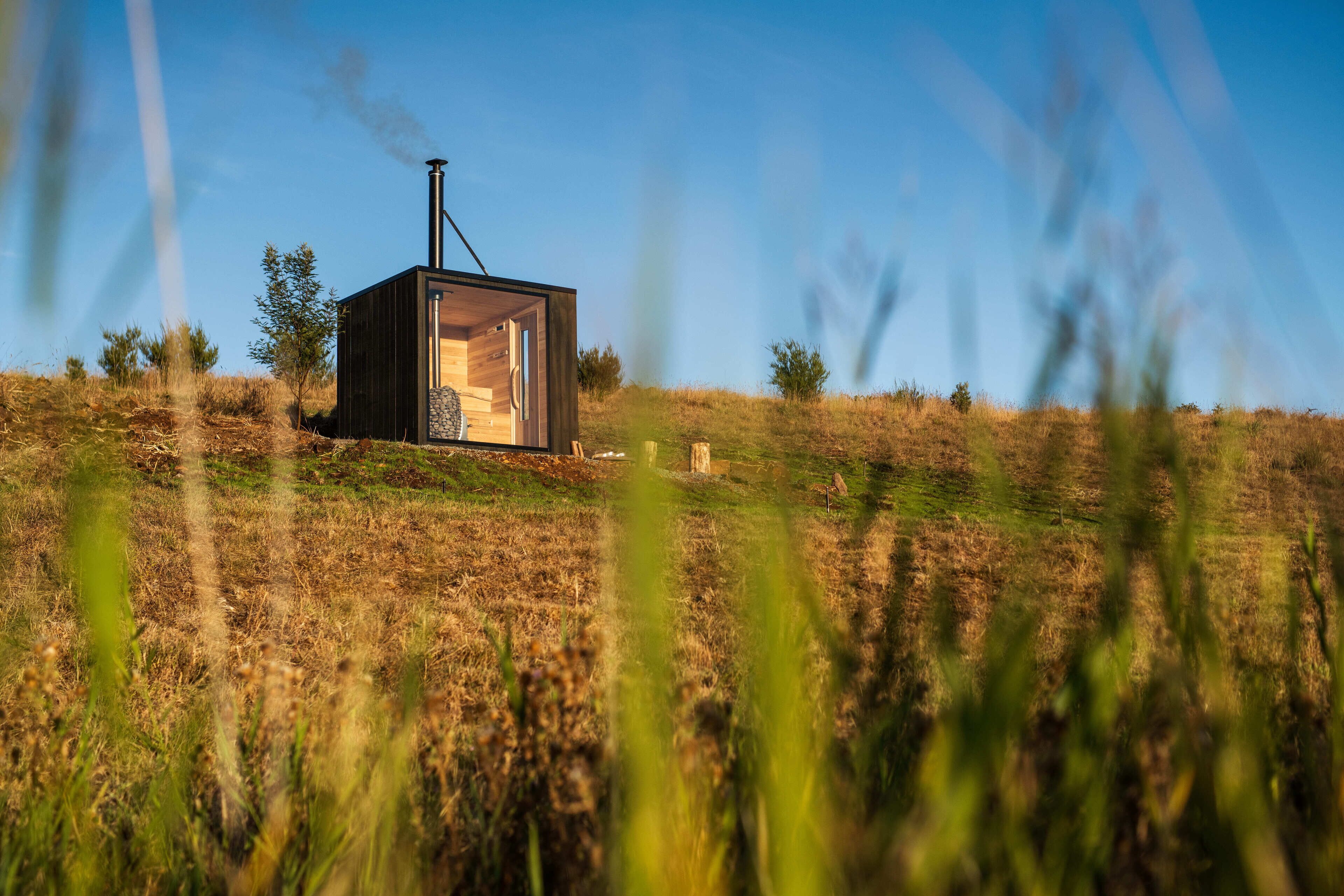 Una cabaña moderna y compacta con interior visible, ubicada en una colina cubierta de hierba bajo un cielo despejado.