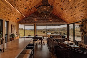 Una habitación amplia con techo de madera, un gran candelabro, una zona de comedor y vistas a un paisaje a través de amplios ventanales.