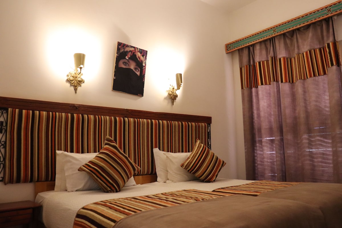 A hotel room with a striped headboard, two wall sconces, a bed with striped bedding, and a large window with curtains.