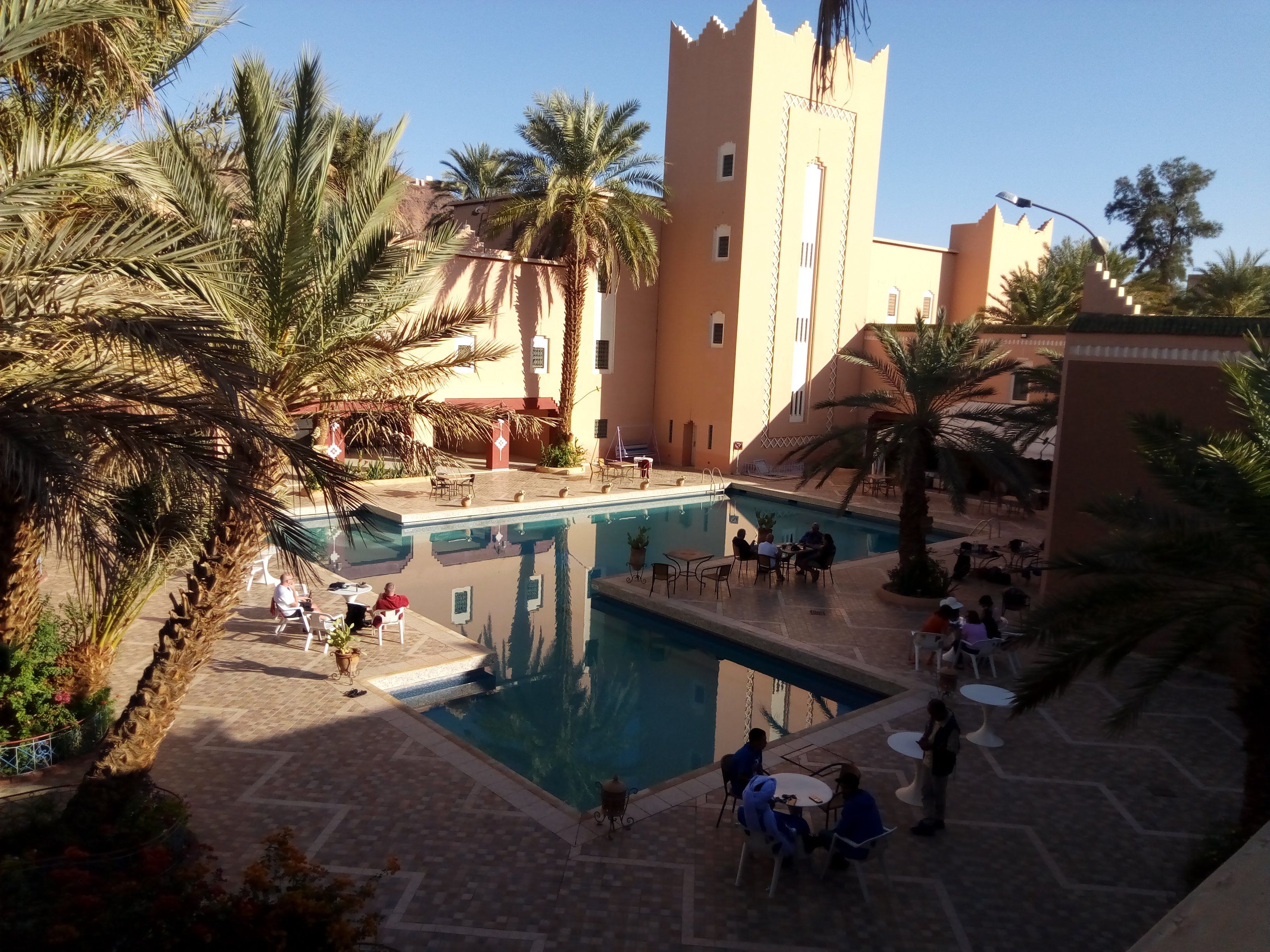 A pool area with palm trees, a castle-like building, and people around tables.