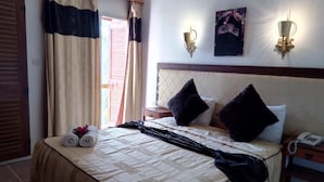 Una cama bien tendida con una colcha beige y negra, dos almohadas negras y una toalla enrollada al lado. Hay una mesita de noche con un teléfono y una lámpara de pared.