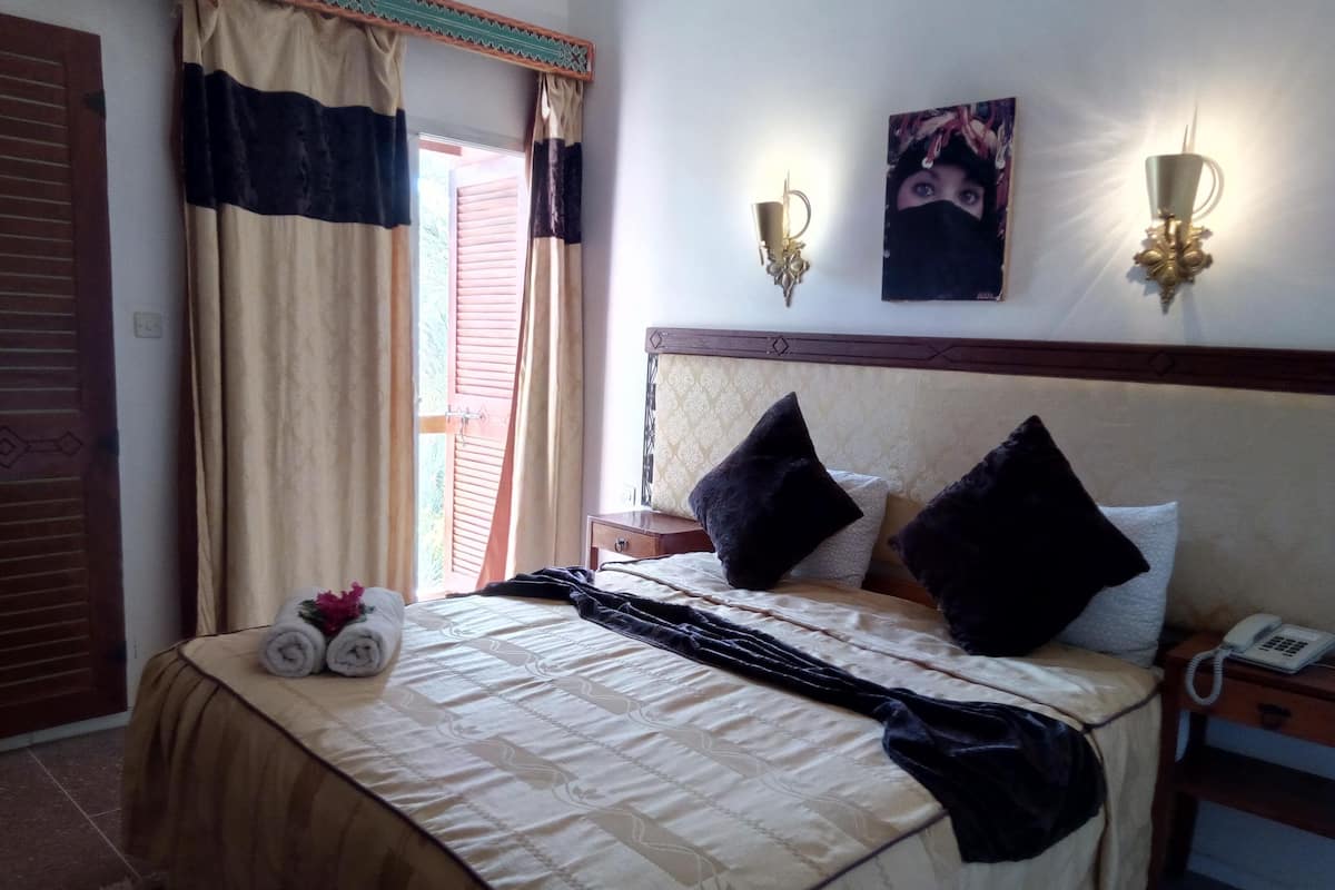 Una cama bien tendida con una colcha beige y negra, dos almohadas negras y una toalla enrollada al lado. Hay una mesita de noche con un teléfono y una lámpara de pared.