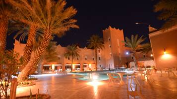 A nighttime view of a courtyard with a pool, palm trees, and a building with multiple balconies.