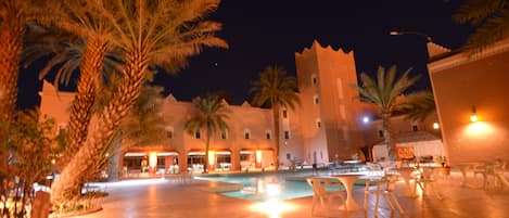 A nighttime view of a courtyard with a pool, palm trees, and a building with multiple balconies.