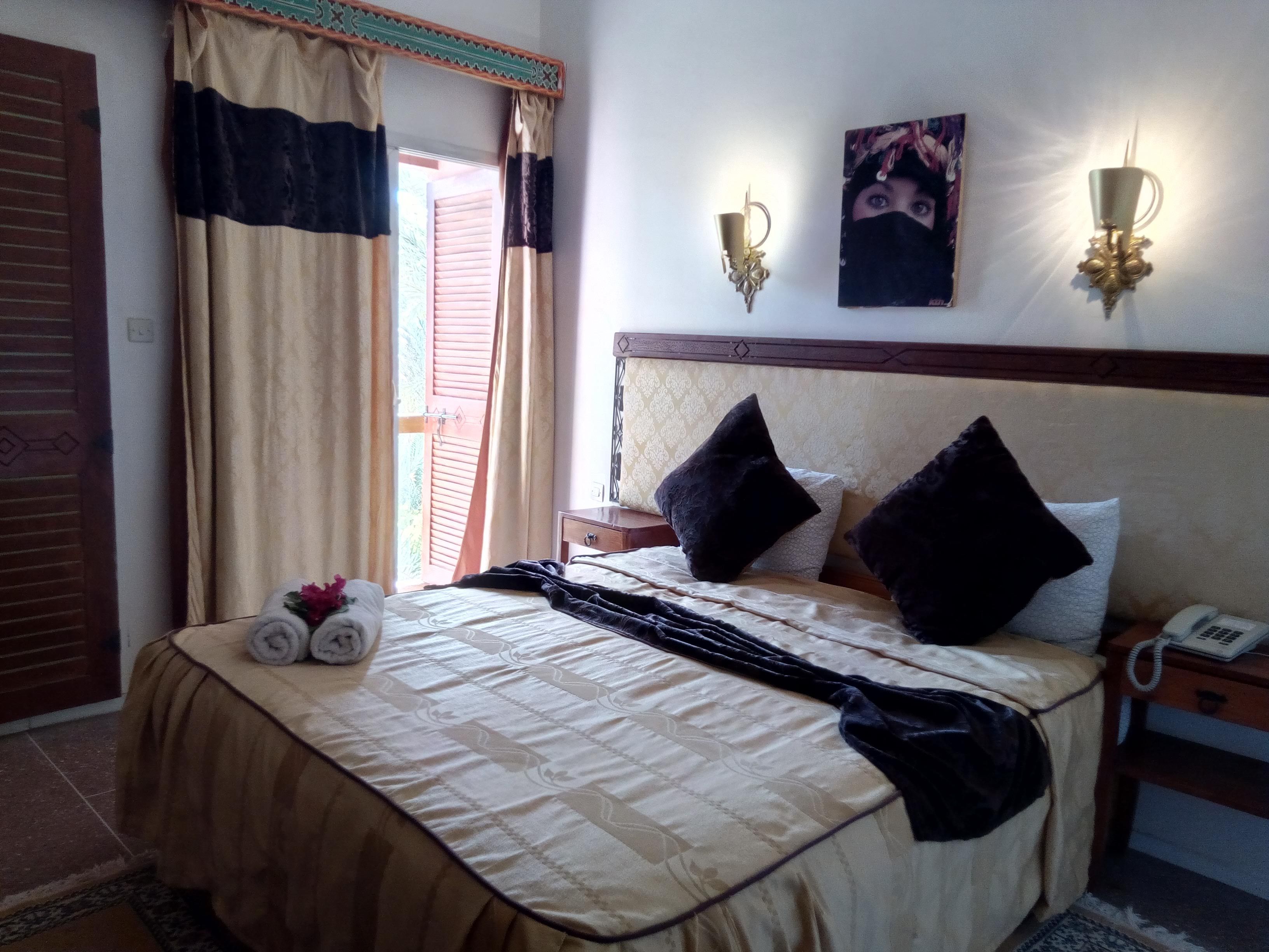 Una cama bien tendida con una colcha beige y negra, dos almohadas negras y una toalla enrollada al lado. Hay una mesita de noche con un teléfono y una lámpara de pared.