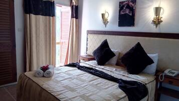 Una cama bien tendida con una colcha beige y negra, dos almohadas negras y una toalla enrollada al lado. Hay una mesita de noche con un teléfono y una lámpara de pared.