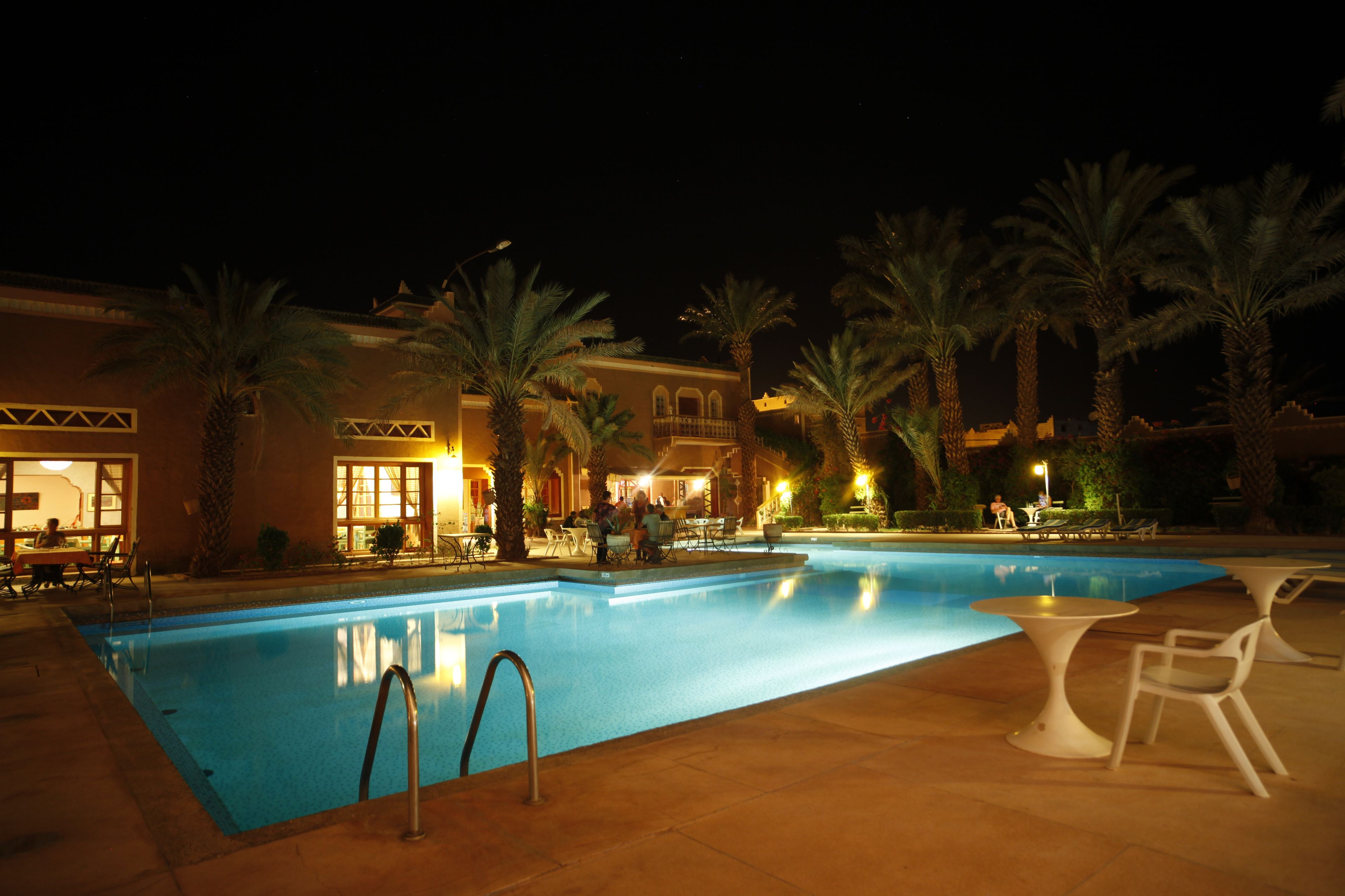 A nighttime swimming pool area with palm trees, outdoor seating, and a building with illuminated windows.