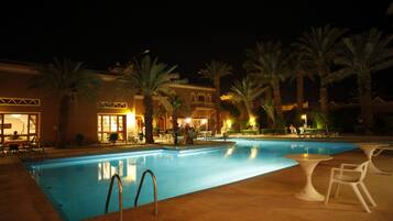 A nighttime swimming pool area with palm trees, outdoor seating, and a building with illuminated windows.