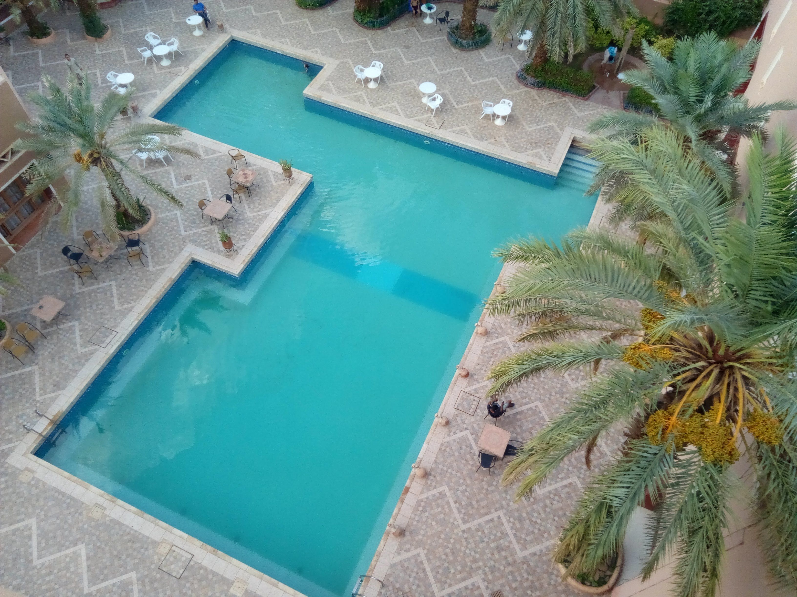 A large rectangular pool with a tiled deck surrounding it, featuring several white tables and chairs.