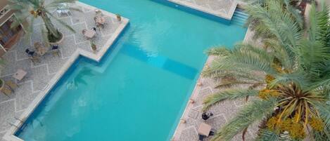 A large rectangular pool with a tiled deck surrounding it, featuring several white tables and chairs.