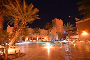 A nighttime view of a courtyard with a pool, palm trees, and a building with multiple balconies. - Le Tinsouline (Zagora)