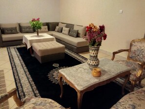 A living room with a sectional sofa, ottoman, a small table with a vase, and a patterned rug. - Le Tinsouline (Zagora)