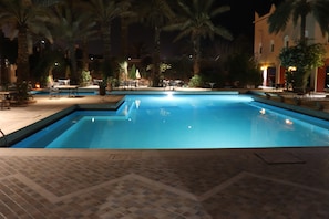 A nighttime swimming pool with tiled flooring and palm trees.