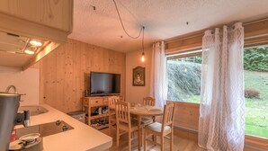 A compact kitchen with wooden cabinets, a built-in stove, a dining table with chairs, a television, and a window with curtains.