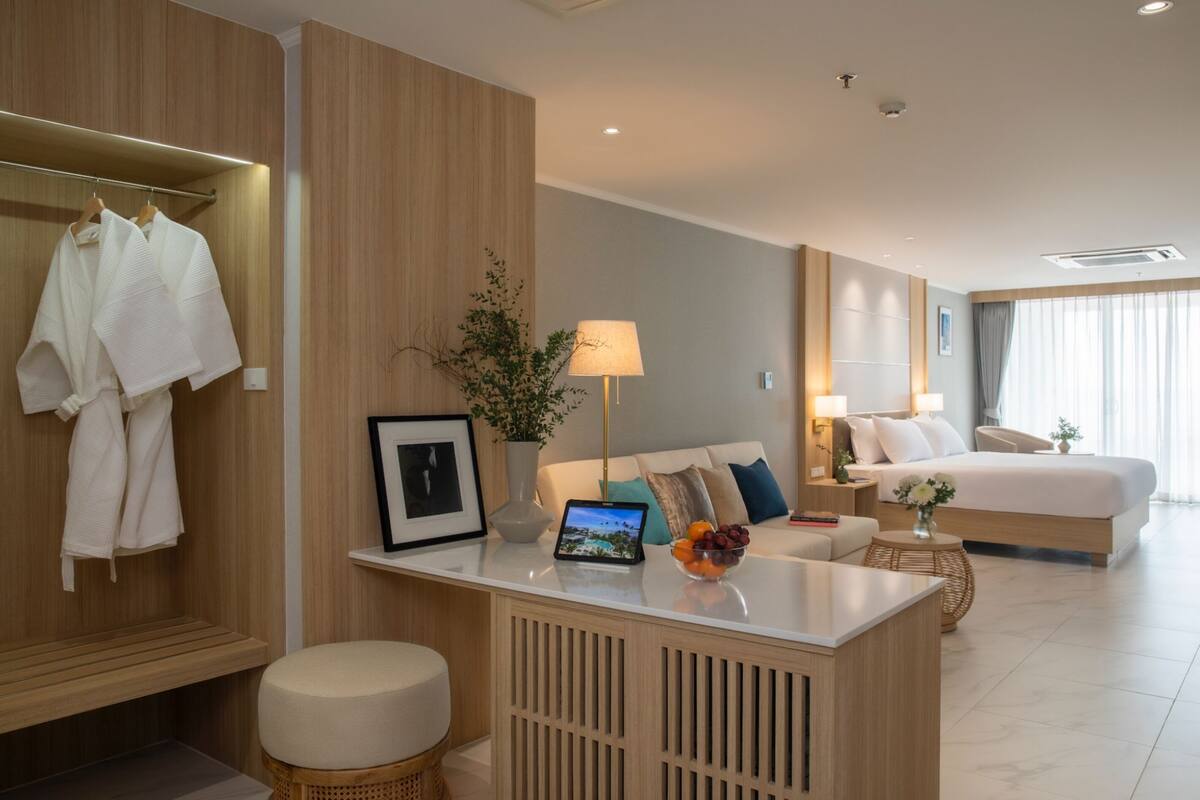 A modern hotel room with a wooden wardrobe, a bedside table with a lamp, a laptop, and a bowl of fruit.