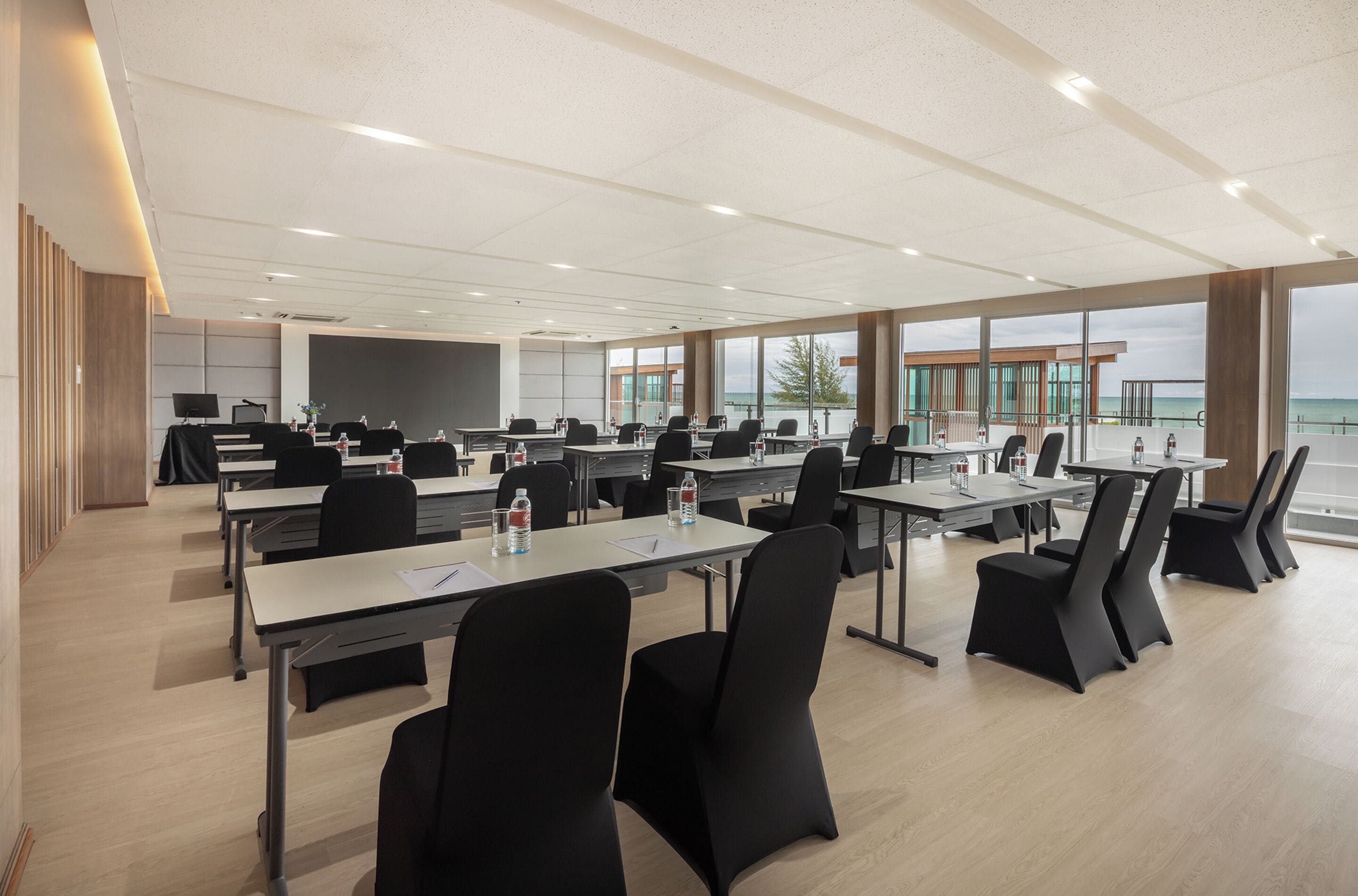 A modern conference room with multiple tables and chairs, large windows offering a view of the sea, and a wooden panel wall on one side.