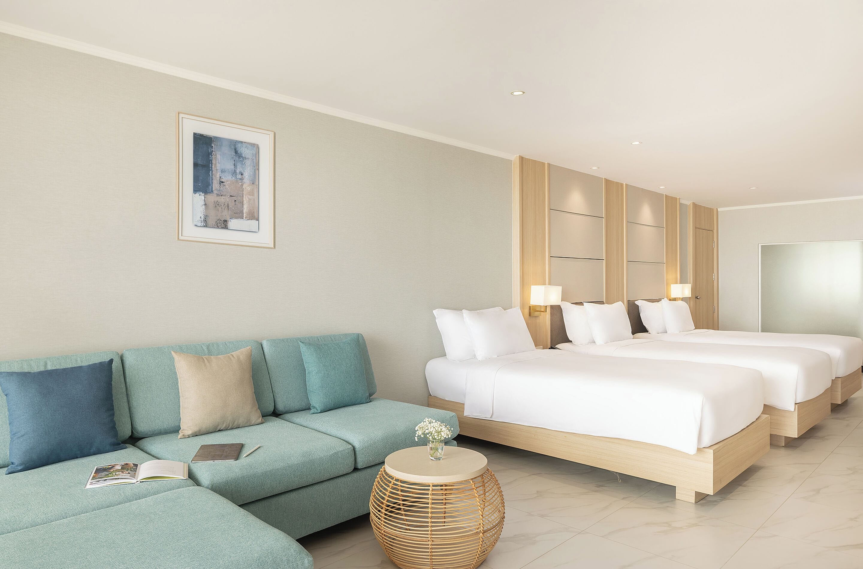 Una habitación de hotel moderna con un sofá de color claro, una mesita auxiliar de mimbre redonda y una cama grande con dos almohadas.